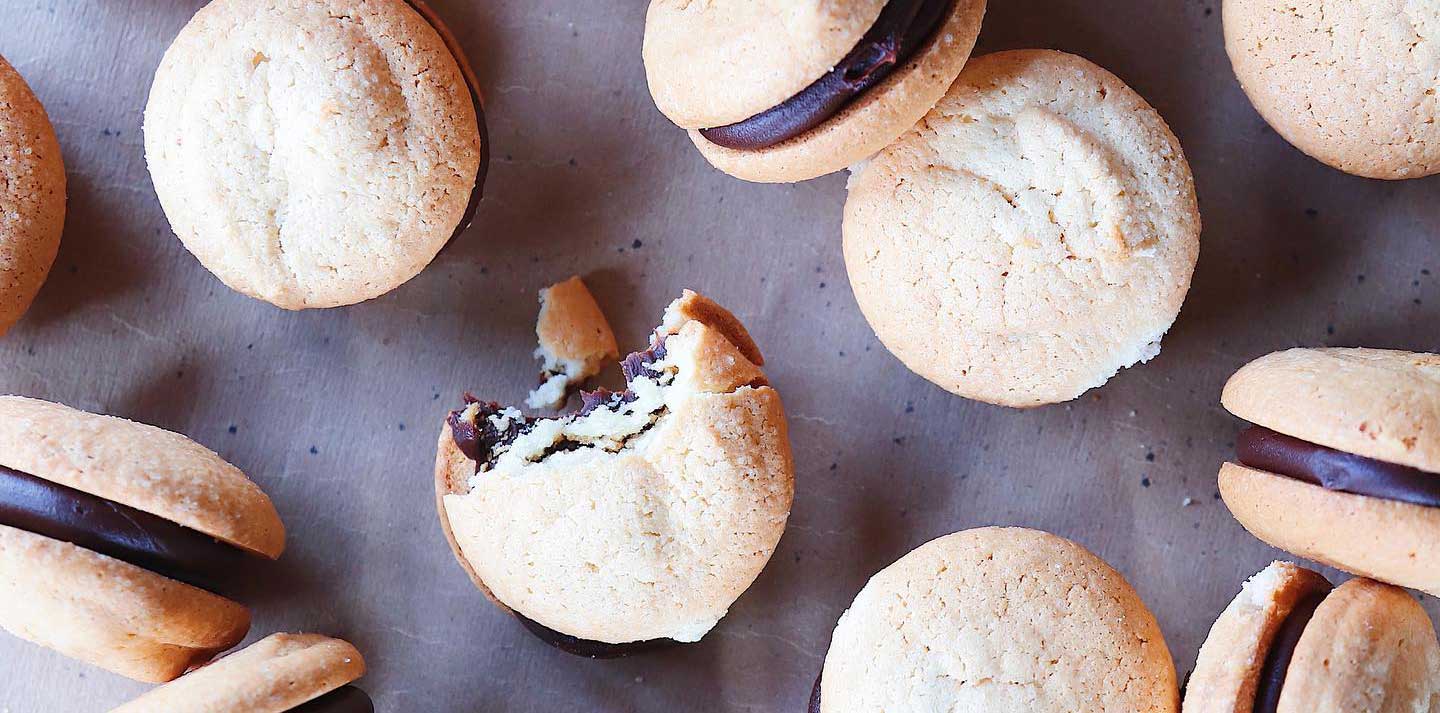 Mini almond macaroon cookies filled with chocolate spread out on a table randomly. One has a bite taken out of it and crumbs are on the table.