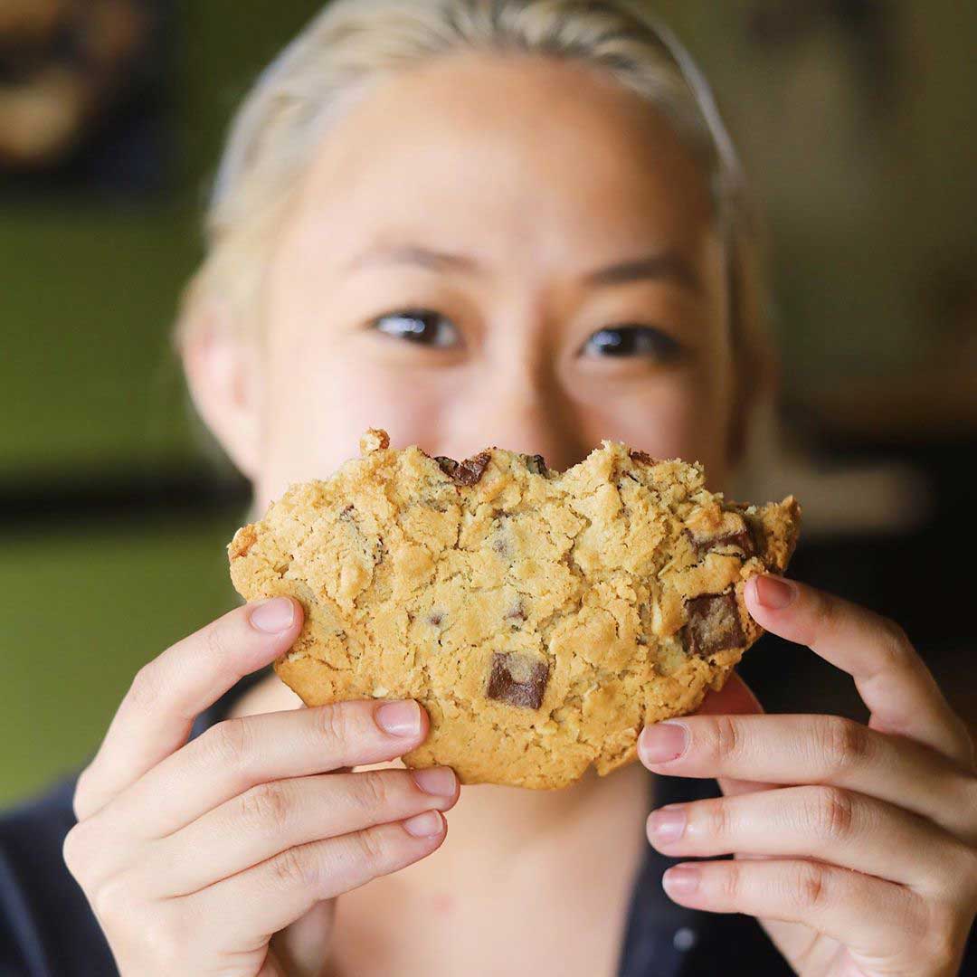 Woman smiling for the camera and holding half eaten giant cookie up to block the bottom half of her face